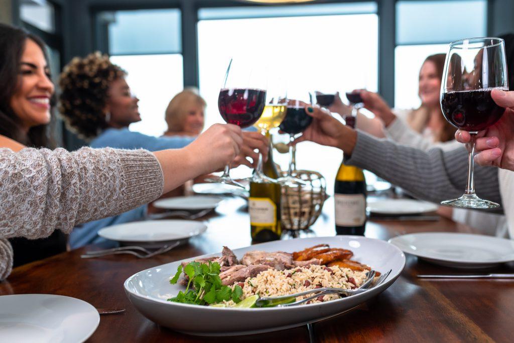 People toasting wine glasses and smiling around a Thanksgiving table with a gourmet meal in the foreground