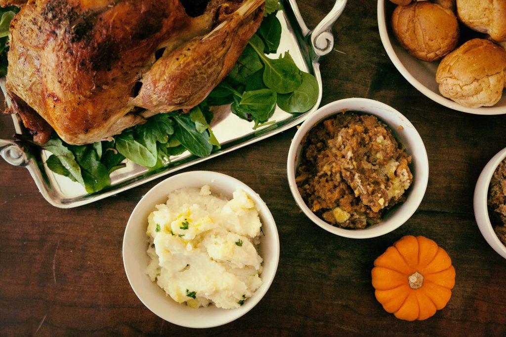 View of a cooked Thanksgiving dinner displayed on a wooden table including turkey, mashed potatoes, stuffing, rolls, and a small orange pumpkin